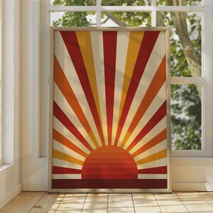 May include: A rectangular framed wall art print featuring a graphic sunburst design. The artwork displays a semi-circular sun at the bottom with alternating rays in shades of deep red, bright orange, and golden yellow against a cream background.