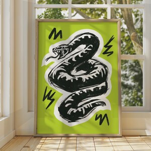May include: A framed wall art print featuring a black ink-style illustration of a coiled, hissing snake with an open mouth and visible tongue. The snake is set against a bright lime green background with black zigzag markings surrounding it.