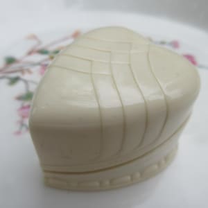 May include: Ivory-colored heart-shaped trinket box with a textured, curved lid. The box has a decorative base and is set against a floral patterned background. The box is likely made of a hard material.