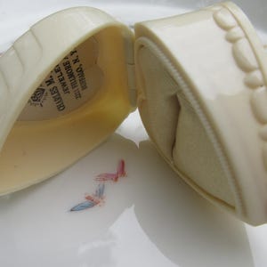 May include: An open, off-white heart-shaped jewelry box. The box has a textured exterior and a soft, beige interior. The text "CHARLES M. JEWELERS" is visible inside the lid. The box is on a white surface with floral accents.