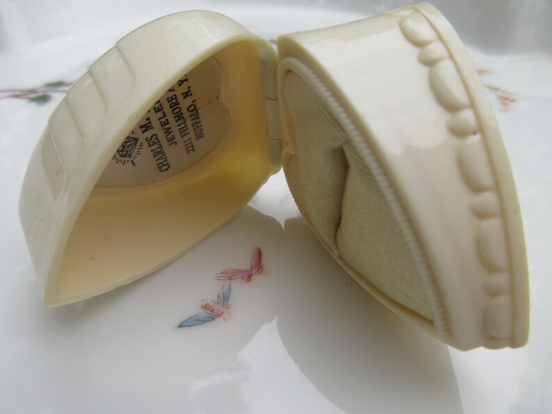 May include: An open, off-white heart-shaped jewelry box. The box has a textured exterior and a soft, beige interior. The text "CHARLES M. JEWELERS" is visible inside the lid. The box is on a white surface with floral accents.