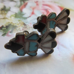 May include: A pair of silver-toned stud earrings with a Native American design. Each earring features a central shape with black, red, and turquoise inlays, and a fan-shaped design with white and black segments. The earrings are set against a floral background.
