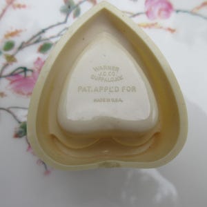 May include: A heart-shaped, off-white trinket dish with a central heart-shaped indentation. The dish has the text "WARNER J.C. CO. BUFFALO, NY. PAT. APPL'D FOR. MADE IN U.S.A." The dish is on a floral patterned surface.