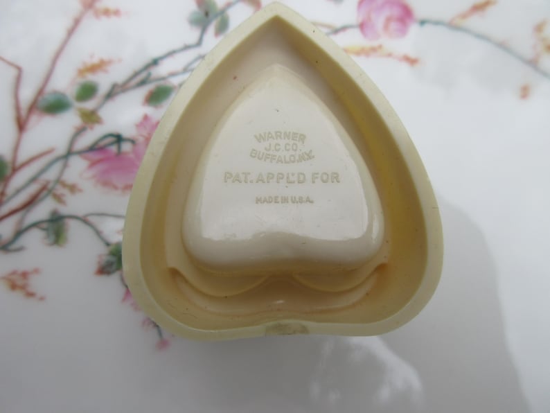 May include: A heart-shaped, off-white trinket dish with a central heart-shaped indentation. The dish has the text "WARNER J.C. CO. BUFFALO, NY. PAT. APPL'D FOR. MADE IN U.S.A." The dish is on a floral patterned surface.