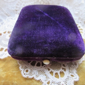 May include: A vintage, rectangular jewelry box with a deep purple velvet exterior. The box has a rounded top and a small, round, off-white clasp. It rests on a white lace doily over a yellow surface.