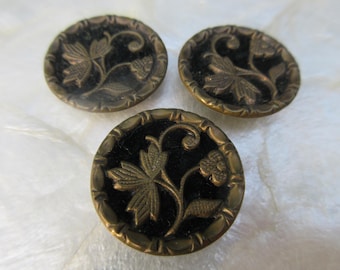 May include: Three antique brass buttons with a black center and a floral design. The buttons are round with a detailed leaf and flower pattern. The buttons are approximately 1 inch in diameter.