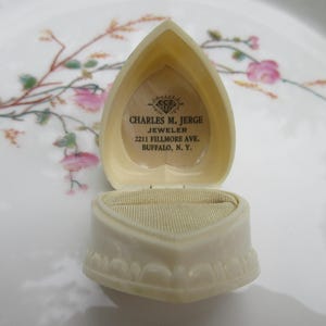 May include: An open, heart-shaped, off-white jewelry box with a textured exterior. The interior features a beige velvet lining and a printed label with the text "CHARLES M. JERGE JEWELER 2211 FILLMORE AVE. BUFFALO, N.Y."
