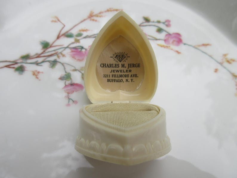 May include: An open, heart-shaped, off-white jewelry box with a textured exterior. The interior features a beige velvet lining and a printed label with the text "CHARLES M. JERGE JEWELER 2211 FILLMORE AVE. BUFFALO, N.Y."