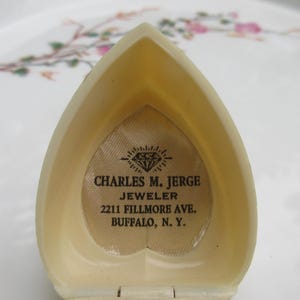 May include: Open, heart-shaped jewelry box in a cream color. The interior features a diamond-shaped logo and the text "CHARLES M. JERGE JEWELER 2211 FILLMORE AVE. BUFFALO, N. Y.". The box is open, revealing the interior.