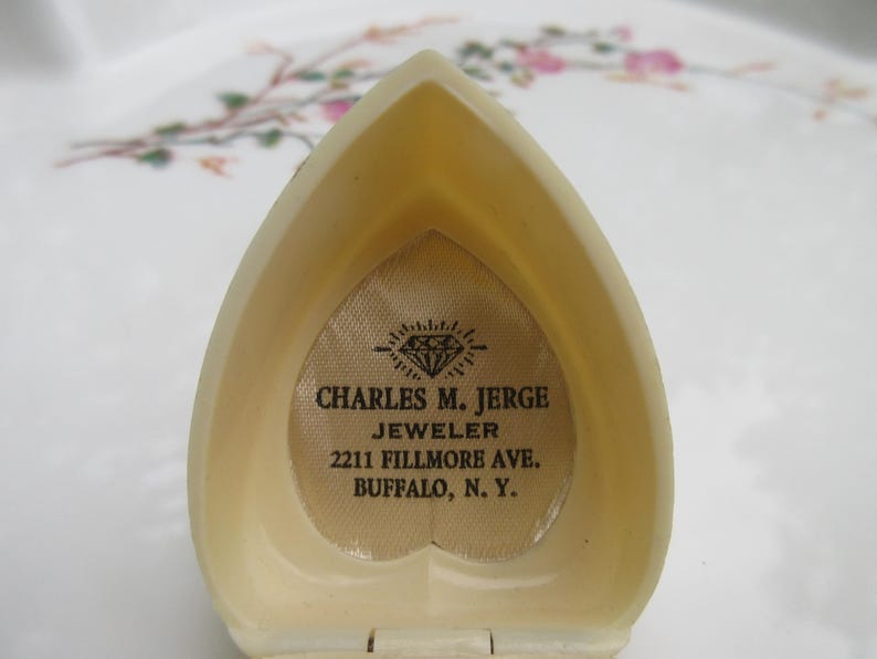 May include: Open, heart-shaped jewelry box in a cream color. The interior features a diamond-shaped logo and the text "CHARLES M. JERGE JEWELER 2211 FILLMORE AVE. BUFFALO, N. Y.". The box is open, revealing the interior.