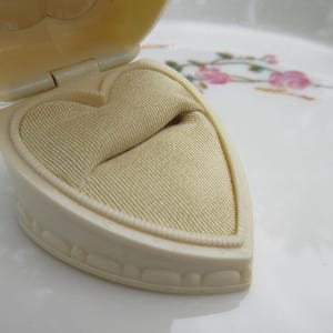 May include: An open, heart-shaped jewelry box in cream color. The interior features a beige, textured fabric cushion. The box is on a white surface with floral patterns.
