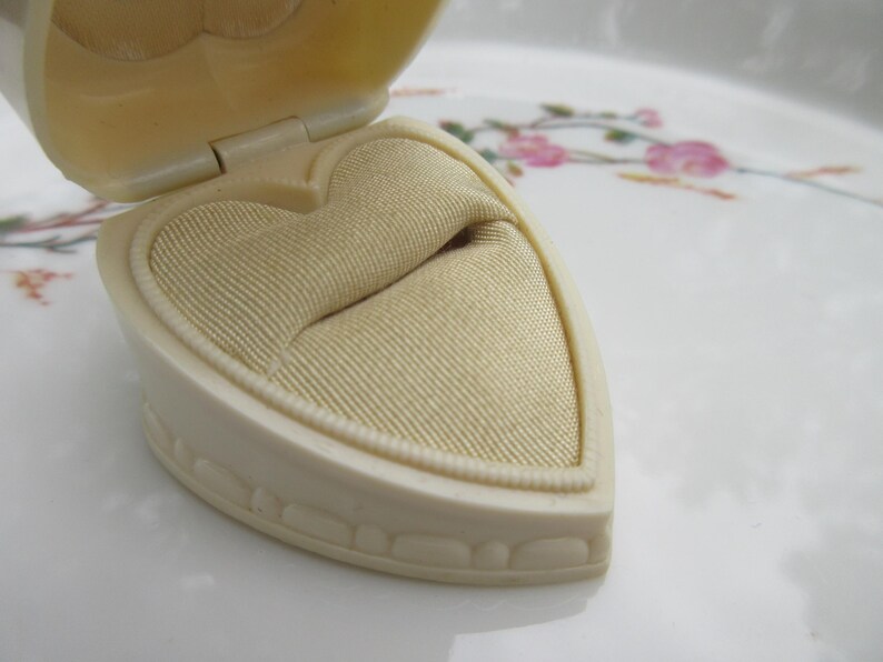 May include: An open, heart-shaped jewelry box in cream color. The interior features a beige, textured fabric cushion. The box is on a white surface with floral patterns.