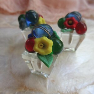 May include: Three clear glass containers, each filled with colorful glass fruit and flowers. The arrangement includes blue, red, green, and yellow elements, with a focus on the vibrant details of the miniature sculptures.