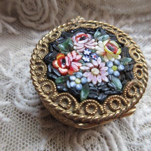 May include: A small, round, antique jewelry box with a mosaic floral design. The mosaic features red, pink, yellow, and blue flowers on a black background. The box has a gold-colored metal frame with a rope-like border.