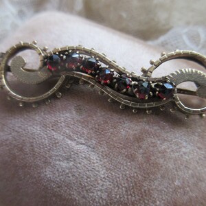 May include: A gold-toned brooch with a delicate, swirling design set with red garnet stones.
