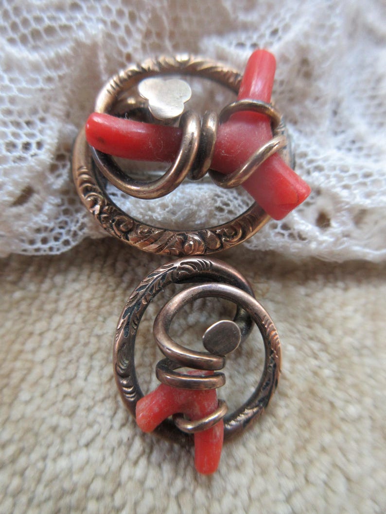 Puede incluir: Un par de pendientes antiguos con un dise&ntilde;o circular. Los pendientes est&aacute;n hechos de metal dorado y presentan una pieza de coral rojo envuelta en espiral. El metal est&aacute; grabado con un patr&oacute;n floral.