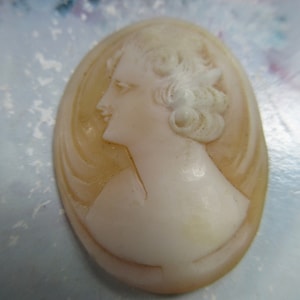 May include: A white cameo featuring a woman's profile with a detailed hairstyle. The cameo is oval shaped and has a light brown background.