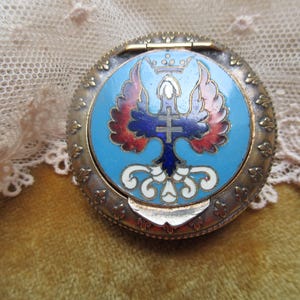 May include: A round, antique metal compact with a hinged lid. The lid features a blue enamel design with a cross and eagle emblem, accented with red, white, and blue details. The compact has a decorative, ornate border.
