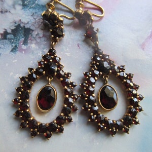 May include: A pair of antique earrings with a gold setting and a large oval-shaped garnet gemstone surrounded by smaller garnets. The earrings have a clip-on closure.
