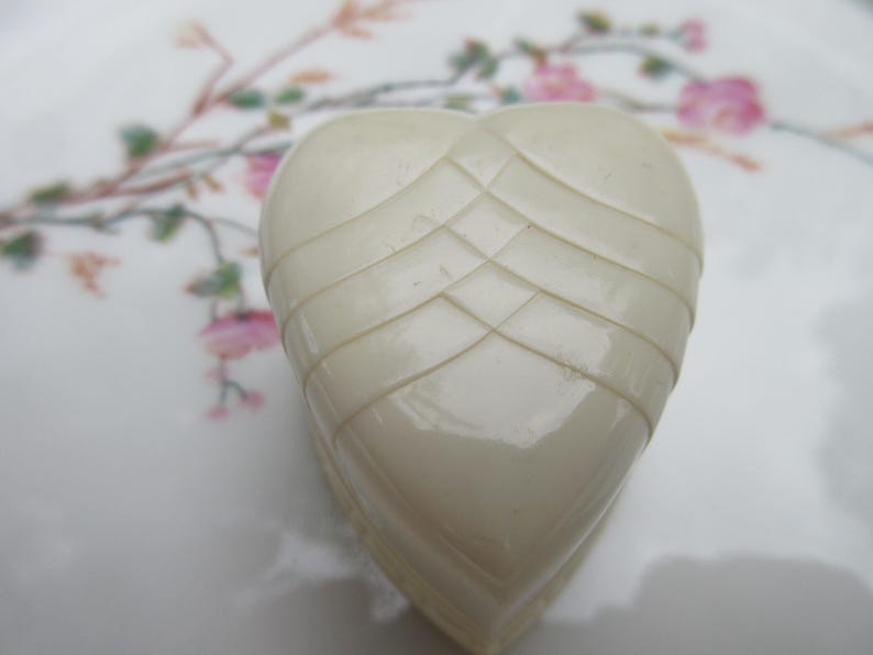 May include: A heart-shaped, off-white decorative object with a textured, layered design. The object is set against a backdrop of a white surface with a floral pattern in pink and green.