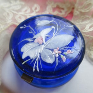 May include: A cobalt blue glass trinket box with a hinged lid. The lid is decorated with a white floral design with pink accents.