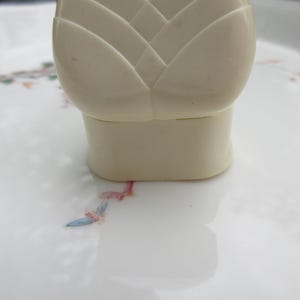 May include: A close-up of a cream-colored bottle stopper with a floral design. The stopper has a geometric, layered top and a cylindrical base. The stopper sits on a white surface with a delicate floral pattern in pink, blue, and green.