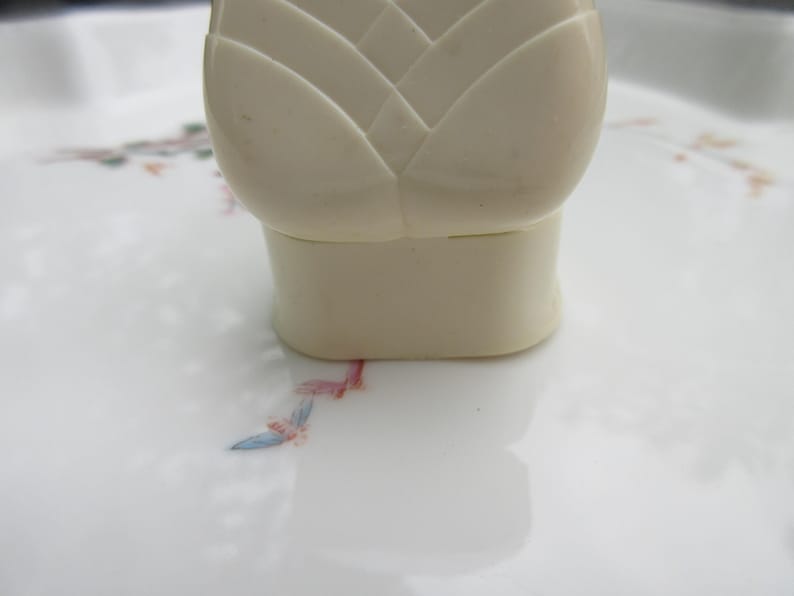 May include: A close-up of a cream-colored bottle stopper with a floral design. The stopper has a geometric, layered top and a cylindrical base. The stopper sits on a white surface with a delicate floral pattern in pink, blue, and green.