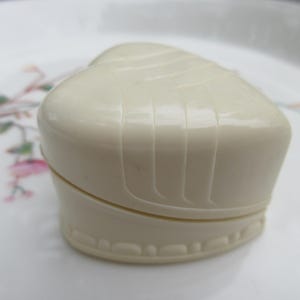May include: A heart-shaped, off-white trinket box with a ridged top and a decorative base. The box is closed, showing a seam where the lid meets the base. The box is sitting on a white plate with floral designs.
