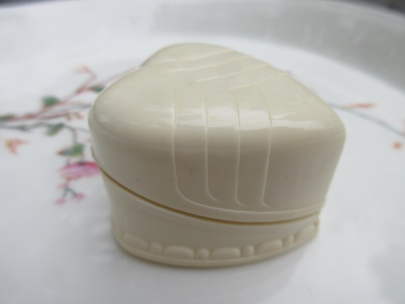 May include: A heart-shaped, off-white trinket box with a ridged top and a decorative base. The box is closed, showing a seam where the lid meets the base. The box is sitting on a white plate with floral designs.