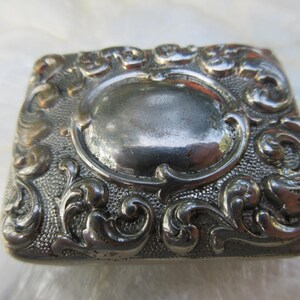 May include: A silver-toned metal box with an ornate, floral design. The box has a raised, oval-shaped center.