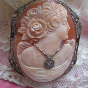 May include: A cameo brooch featuring a carved portrait of a woman with a rose in her hair. The cameo is set in a silver filigree frame and has a small diamond pendant hanging from a chain.