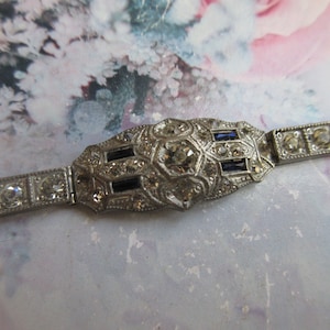May include: A silver bracelet with a large, ornate centerpiece featuring a clear rhinestone surrounded by smaller rhinestones and blue stones. The bracelet has a simple, straight design.
