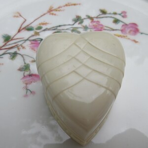 May include: Ivory-colored heart-shaped trinket box with a woven design on the top. The box sits on a white plate decorated with pink floral and green leaf patterns.
