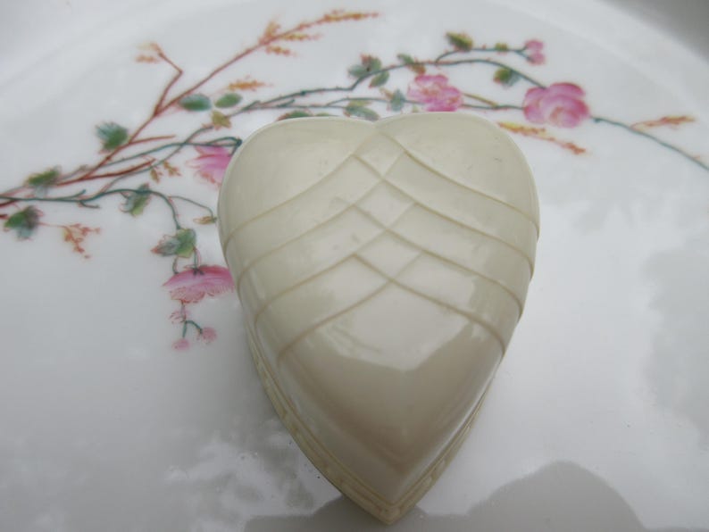 May include: Ivory-colored heart-shaped trinket box with a woven design on the top. The box sits on a white plate decorated with pink floral and green leaf patterns.