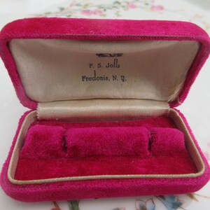 May include: A rectangular, open jewelry box covered in vibrant fuchsia velvet. The interior features a cream-colored satin lining with the printed text F. S. Jolls Fredonia, N. Y. and a fuchsia velvet insert with three slots for holding jewelry.