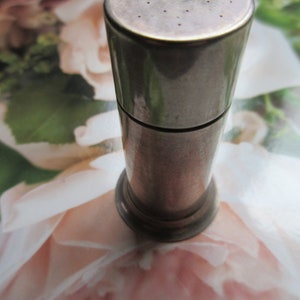 May include: A silver metal pepper shaker with a round top and a small hole in the center. The shaker has a smooth, shiny surface.