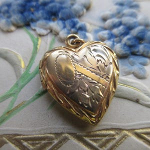 May include: A gold heart-shaped locket with intricate floral and leaf engravings. The locket has a small loop at the top for a chain. The background features blue floral accents and a white surface with gold trim.