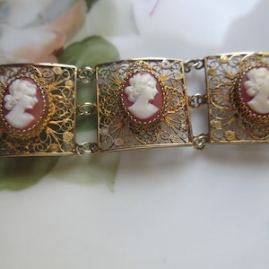 May include: A gold-tone metal bracelet featuring three rectangular filigree links, each centered with an oval-shaped cameo. The cameos display a white profile portrait of a person against a reddish-brown background, connected by small metal links.