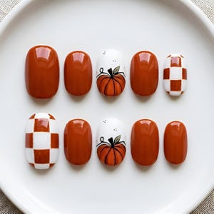 May include: A set of ten press-on nails with a glossy finish. The design features solid burnt orange nails, white nails with hand-painted orange pumpkins and black stems, and nails with a white and burnt orange checkered pattern arranged in two rows.