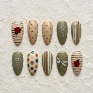 May include: Ten almond-shaped press-on nails featuring various designs including a red ladybug on beige stripes, brown polka dots on beige, a sun motif on olive green, vertical stripes, and a dragonfly illustration on an olive green background.