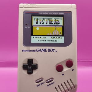 Può includere: Una console portatile Nintendo Game Boy grigia, con il gioco Tetris sullo schermo. Il dispositivo ha un pad direzionale nero, pulsanti A e B rossi e pulsanti select e start. Lo schermo mostra il logo Tetris e il gameplay.