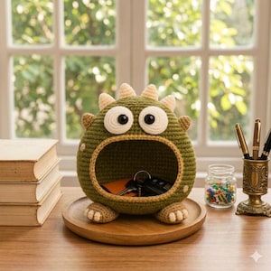 May include: A round, olive green crocheted monster organiser with a wide, open mouth for storage. It features two large white eyes with black pupils, small beige horns on top, and beige feet. The interior holds a set of metal keys on a ring.