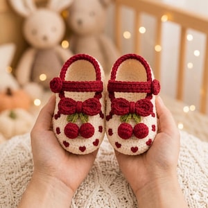 Puede incluir: Un par de zapatos de bebé tejidos a ganchillo en color crema con ribete rojo oscuro, una correa a juego con botón y un lazo decorativo. Los zapatos están adornados con pequeños corazones rojos y un par de cerezas rojas colgantes.