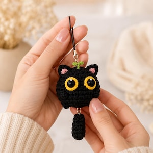 May include: A black crocheted cat charm featuring large yellow-rimmed eyes, small pink inner ears, and a tiny green sprout on top. The cat has a dangling black crocheted tail attached by a metal ring and a black cord loop for hanging purposes.