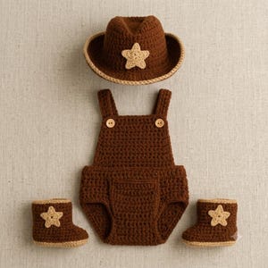 May include: A brown crochet set consisting of a cowboy hat with a beige star, a pair of overalls with two wooden buttons, and two matching booties each featuring a beige star. The items are crafted from dark brown yarn with a textured stitch.