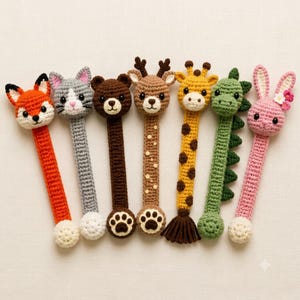 May include: A set of seven crochet animal bookmarks featuring a fox, cat, bear, deer, giraffe, dinosaur, and rabbit. Each bookmark has a long, slender body with a detailed animal head at the top and a small tail or paw-shaped end at the bottom.