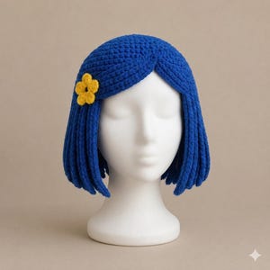 May include: A blue crocheted wig with a bob-length cut and a center part. The wig features a small, bright yellow crocheted flower accessory attached to the side. The yarn has a textured, looped appearance, and the wig is displayed on a white mannequin head.