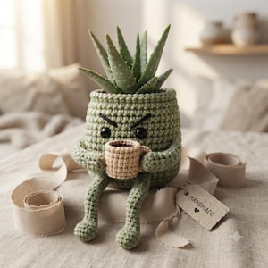 May include: A sage green crocheted planter shaped like a character with an angry expression, holding a small beige mug. The planter contains a green aloe plant with spiky leaves. A small rectangular tag attached by string reads HANDMADE.