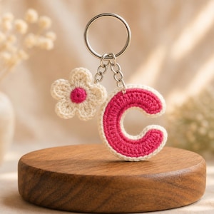 Puede incluir: Un llavero de ganchillo con una letra C y un pequeño dije de flor. La letra C está elaborada con un centro rosa brillante y un borde color crema. La flor tiene pétalos color crema y un centro rosa. Ambos están unidos a una argolla de metal.