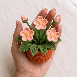 Könnte beinhalten: Eine kleine gehäkelte Topfpflanze mit drei hellrosa Blüten mit gelben Mitten, zwei geschlossenen Blütenknospen und mehreren dunkelgrünen Blättern. Die Pflanze befindet sich in einem runden, strukturierten, terrakottafarbenen Häkeltopf.